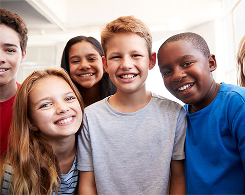 A group of six diverse children smiling and standing close together, looking at the camera indoors.
