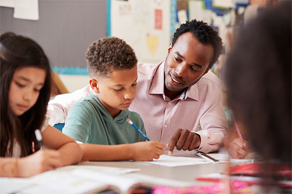 Teacher helping young students with their schoolwork at a table in a classroom setting.