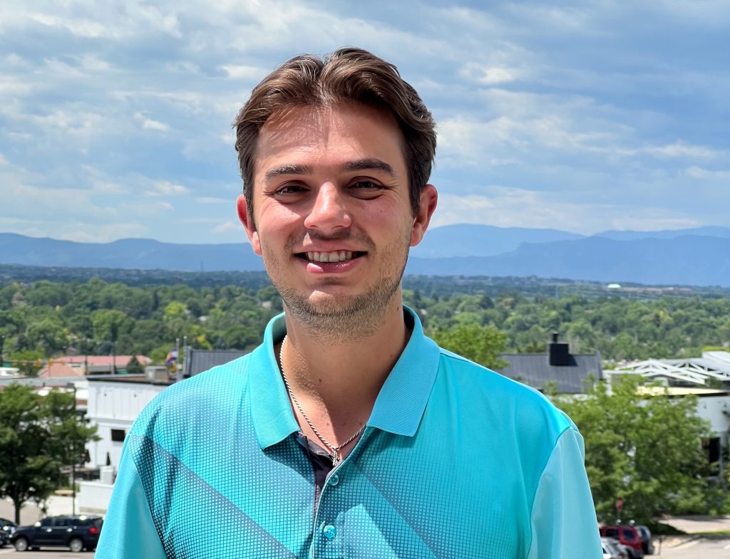 A person in a blue shirt smiles outdoors with a scenic backdrop of trees, buildings, and distant mountains.